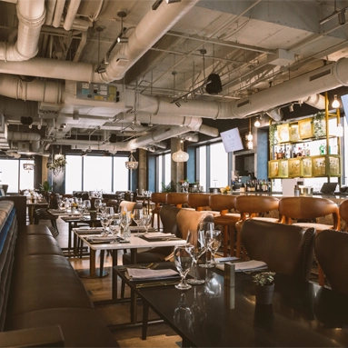 Modern restaurant interior with brown leather chairs, set tables with glassware, large windows letting in natural light, and exposed pipes on the ceiling—perfectly situated near Class A office space San Antonio. The bar area is visible in the background.