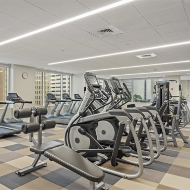 Modern gym in Class A office space San Antonio with large windows, several treadmills, elliptical machines, strength training equipment, and neutral-toned flooring. Brightly lit room offers city building views.