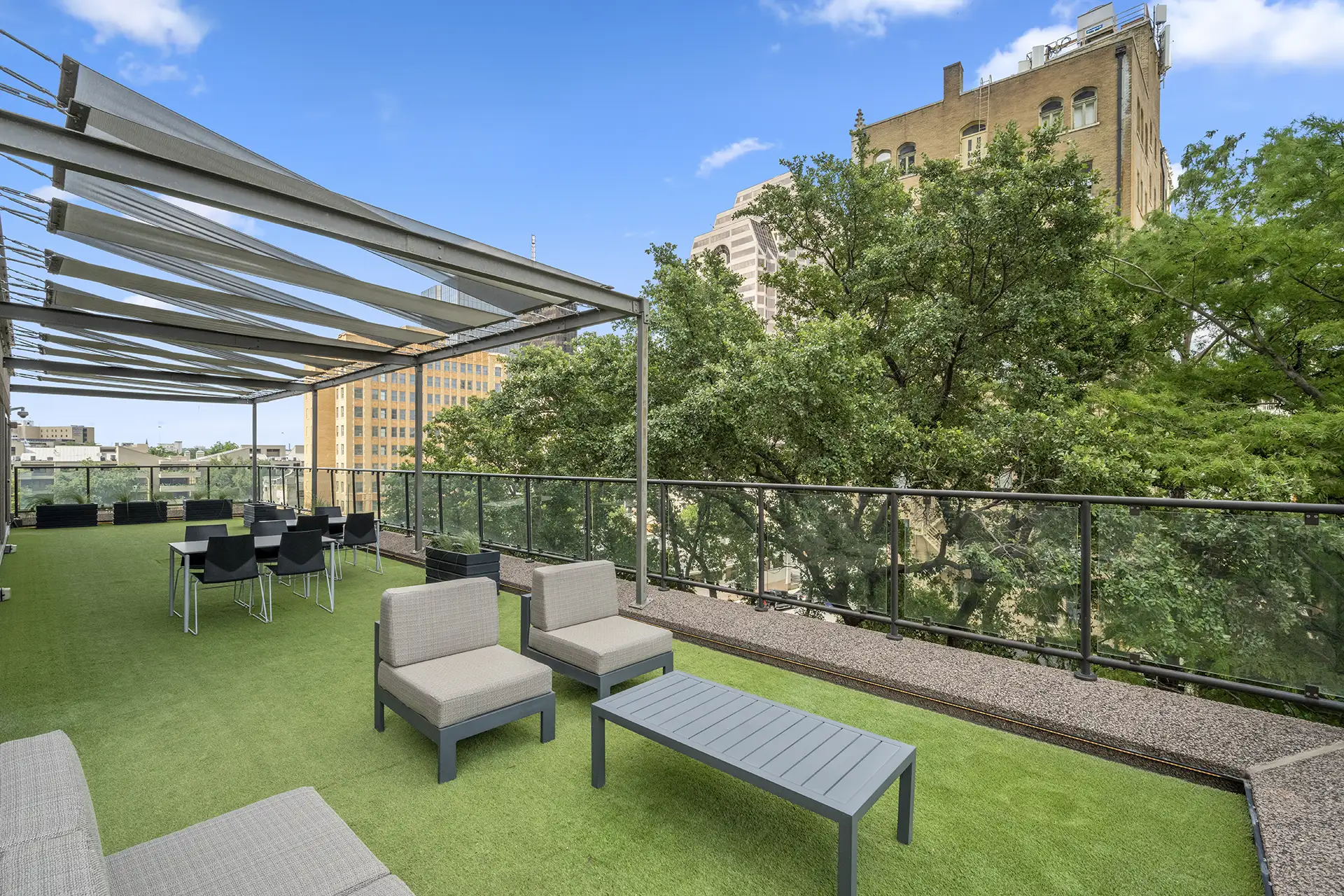 Modern rooftop patio in a Class A office space San Antonio, featuring green artificial grass, lounge chairs, tables, and a pergola. Glass railings offer views of trees and tall city buildings under a clear blue sky.