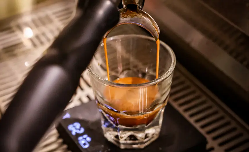 Close-up of espresso being poured from a coffee machine portafilter into a small glass, rich brown crema forming on top. Perfect fuel for your busy morning in Class A office space San Antonio, set on a cafe counter.