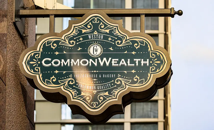 An ornate hanging sign reads Weston CommonWealth Coffeehouse & Bakery, Uncommon Quality in white and gold on a dark green background, mounted on a building with glass windows—steps from Class A office space San Antonio professionals frequent.