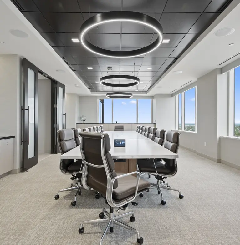 Modern conference room with a long white table surrounded by gray office chairs, large windows with city views, and three circular ceiling lights. The room is bright, spacious, and neatly organized.