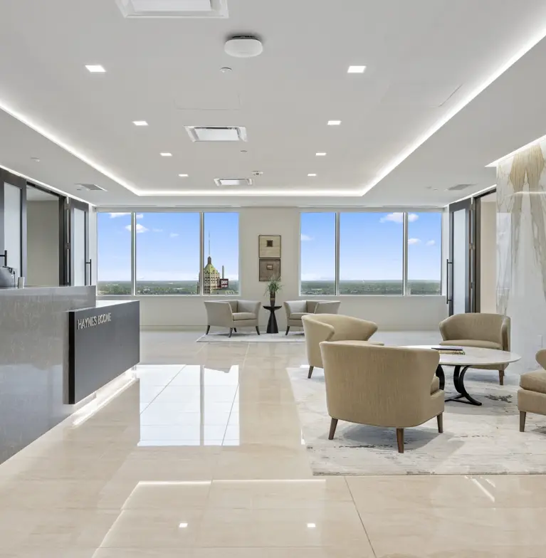 Modern office lobby with beige chairs and tables, a reception desk on the left, large windows with city views, and bright natural light reflecting on glossy floors.