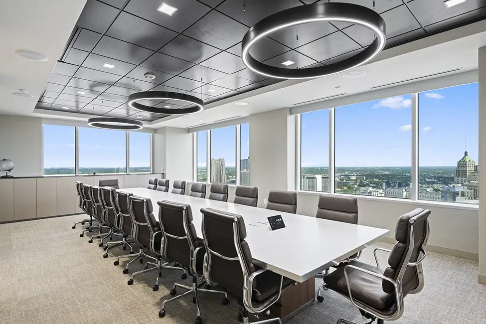 A modern conference room in Class A office space San Antonio, featuring a long white table, numerous office chairs, circular ceiling lights, and large windows with stunning city skyline views.