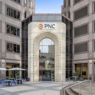Modern office building entrance with large glass windows, a decorative archway, and the PNC logo above the door. Outdoor seating with blue chairs and tables is set up in the foreground, inviting visitors to check suite details on the single post nearby.