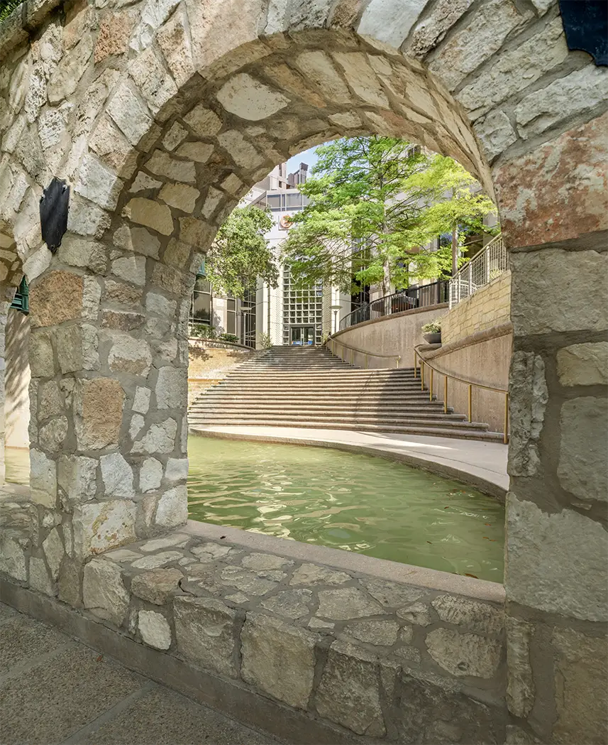 View through a stone archway of a curved staircase leading up from a canal with green water, surrounded by trees and buildings—an inviting scene in an outdoor urban setting near Class A office space San Antonio.