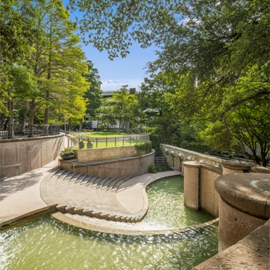 A curved stone staircase descends alongside a flowing waterway, surrounded by lush green trees and open grassy areas under a bright blue sky—an inviting scene near Class A office space San Antonio.
