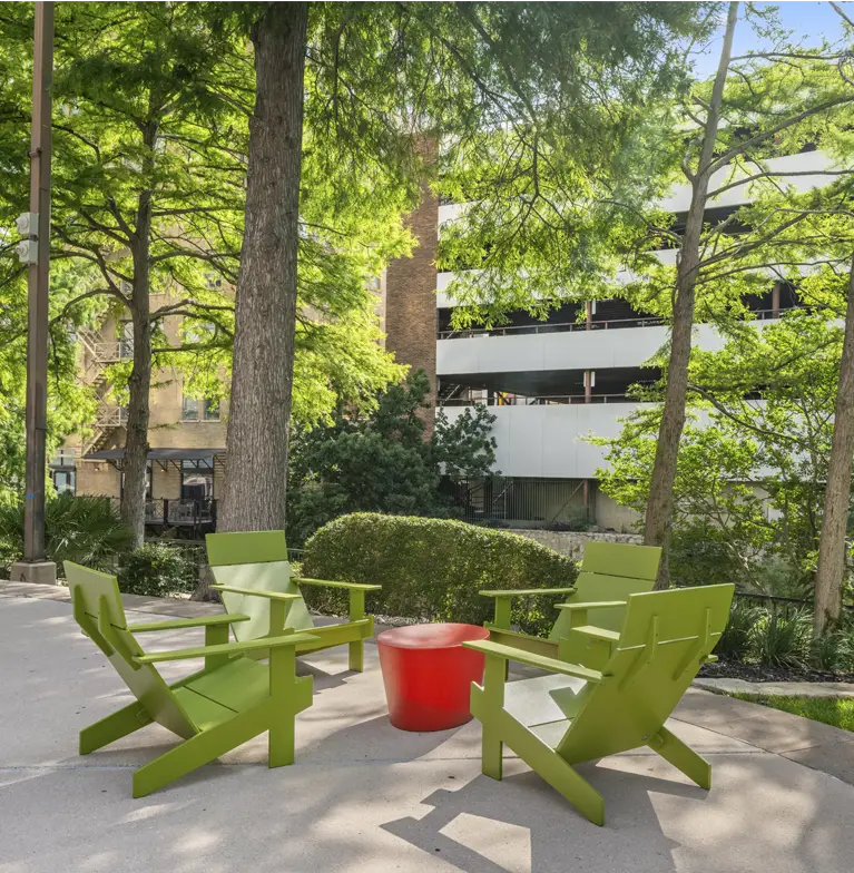 Four modern green chairs arranged around a small red table sit on a paved area under tall leafy trees, with a multi-story parking garage and bushes visible in the background.
