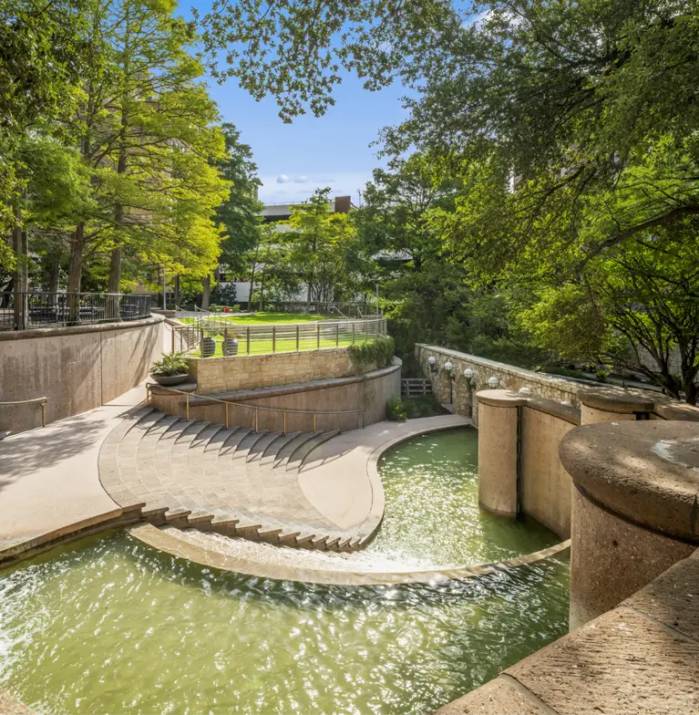 A sunlit outdoor curved, tiered water fountain with cascading water features curved stone steps leading down to a pool of greenish water, surrounded by lush trees and landscaped greenery.