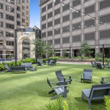 Outdoor courtyard with artificial grass, black Adirondack chairs, and string lights, surrounded by modern tall Class A office space San Antonio buildings. The PNC logo is visible on one building in the background.