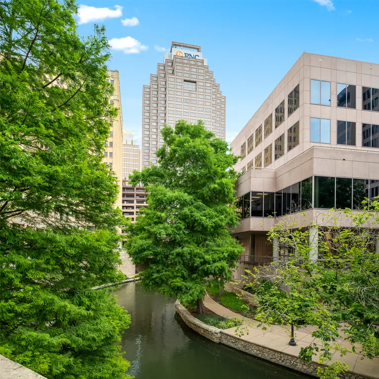 A tree-lined riverwalk curves between modern office buildings in a city, offering views of Class A office space San Antonio, with the PNC Bank building visible in the background under a blue sky with a few clouds.