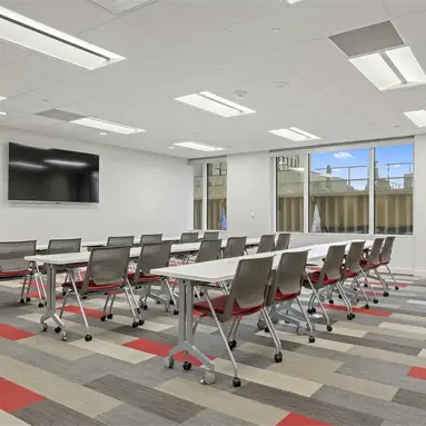 A modern Class A office space San Antonio conference room with rows of tables and chairs on a patterned carpet, a large wall-mounted TV screen, white walls, and expansive windows letting in natural light.