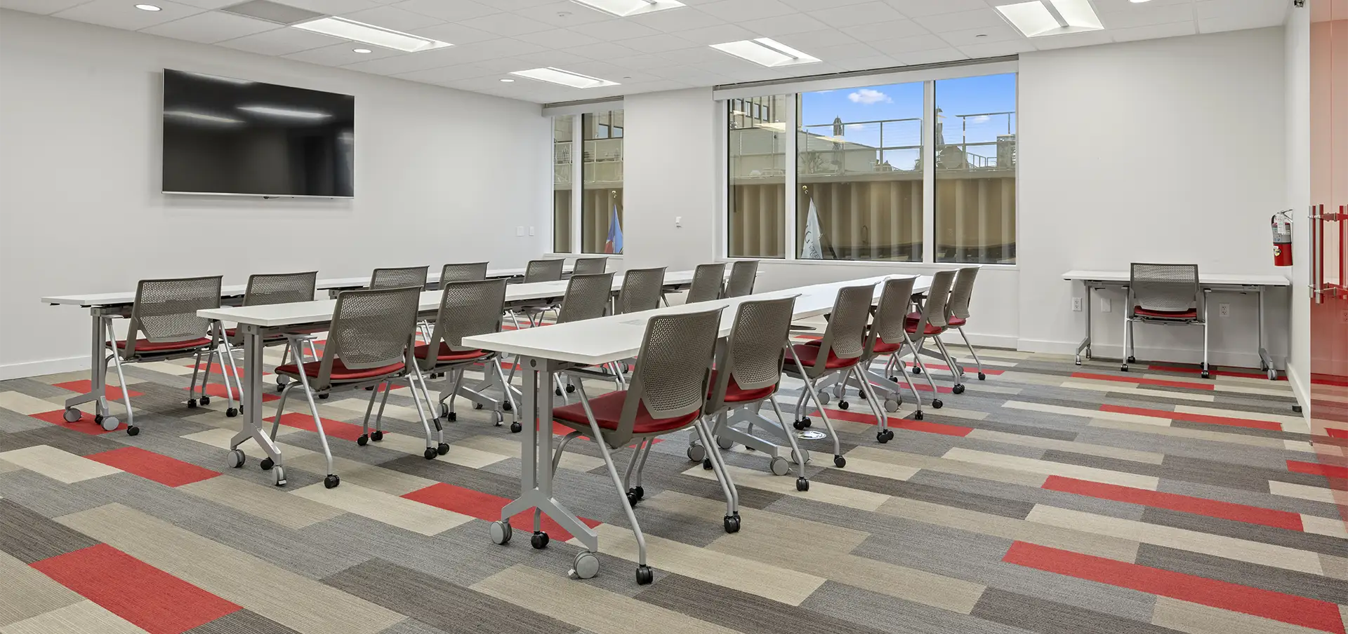 Modern classroom in Class A office space San Antonio with rectangular tables and gray chairs on wheels arranged in rows, facing a large wall-mounted screen. Large windows, white walls, and a colorful carpet in red, gray, and beige patterns complete the look.