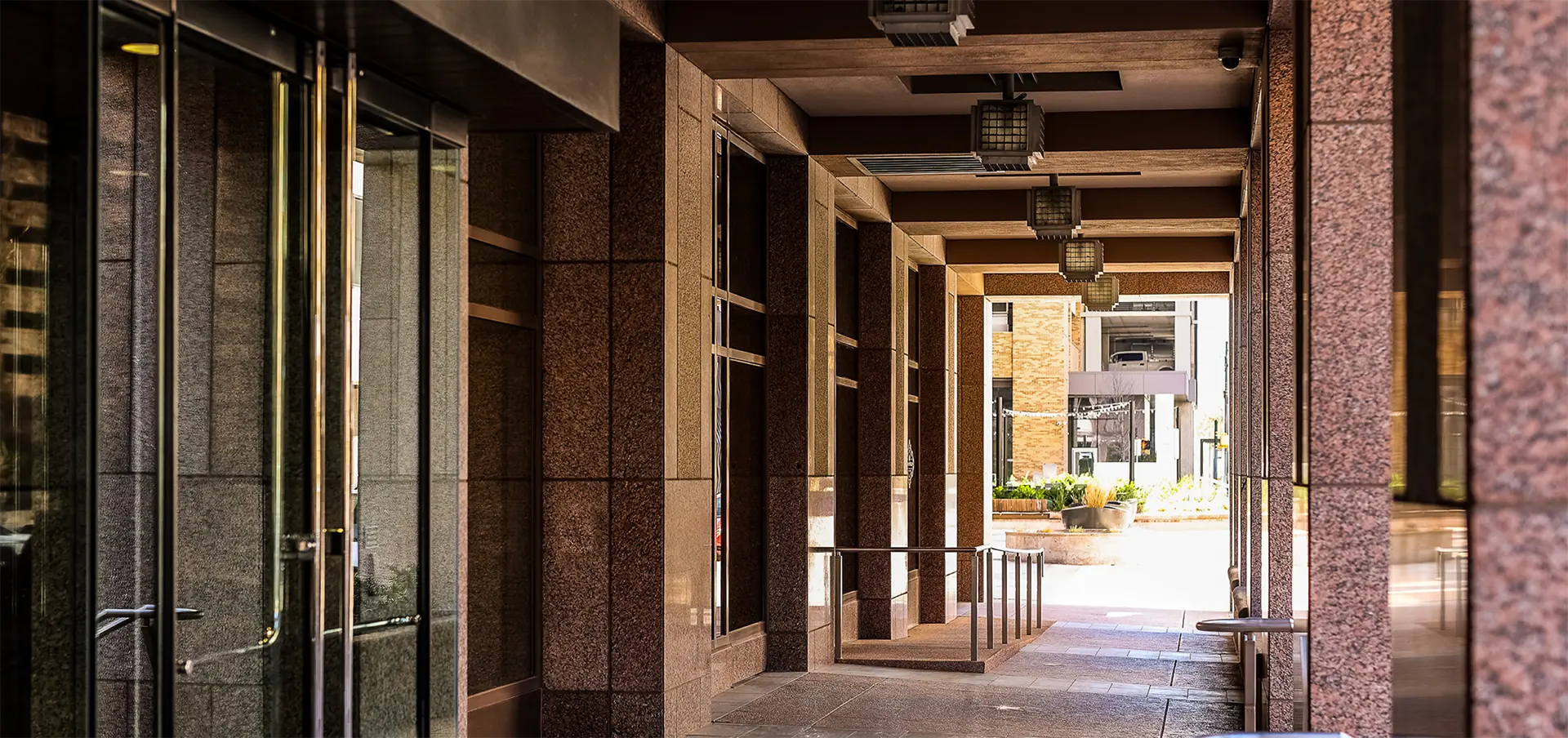 Covered walkway with stone columns and large glass doors on the left, leading toward a sunlit outdoor area with planters—showcasing a welcoming entrance to premier Class A office space in San Antonio.