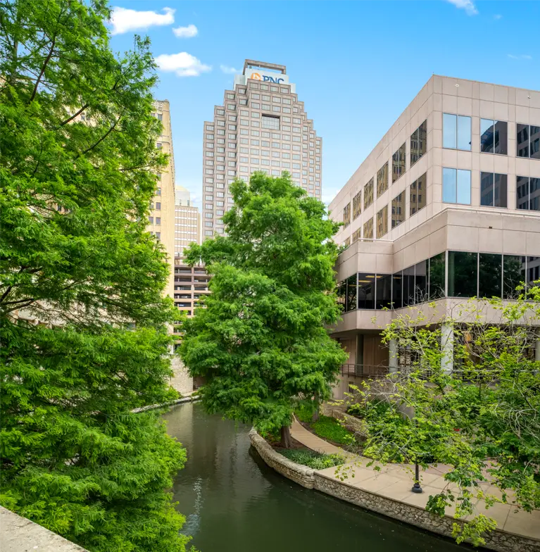 A narrow river winds between modern office buildings and lush green trees, with a tall skyscraper in the background under a bright blue sky with scattered clouds.