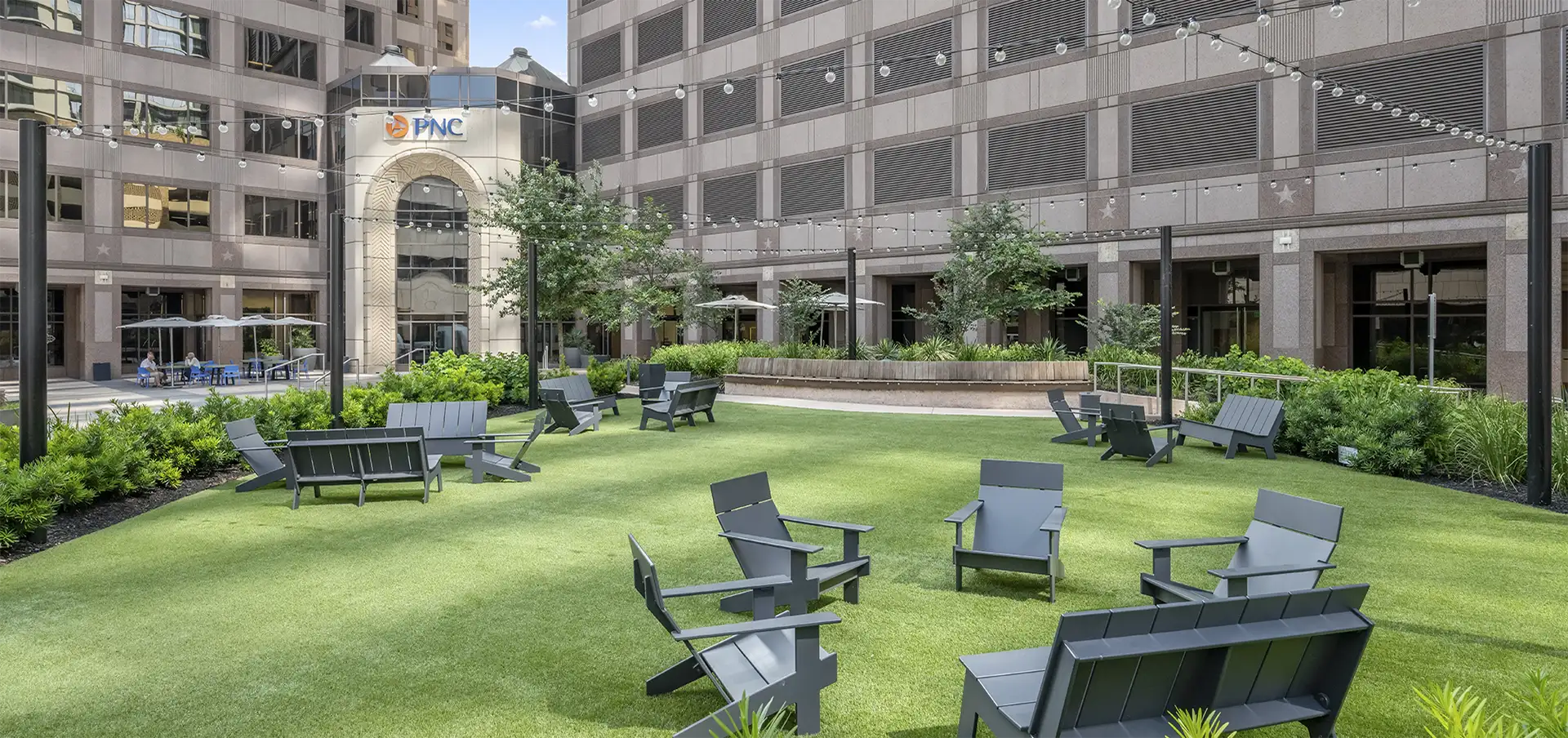 Outdoor courtyard in a city with green artificial grass, black Adirondack chairs in small groups, string lights overhead, lush landscaping, and tall Class A office space San Antonio buildings surrounding the area.